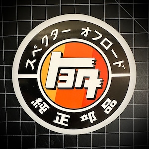 Peut inclure: Autocollant rond avec texte japonais et logo Toyota blanc sur fond orange et jaune dégradé. L'autocollant a une bordure noire et un texte blanc qui se lit "Specter Offroad" et "Genuine Parts".