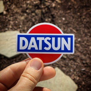 May include: A sticker with the word "DATSUN" in white, set against a blue rectangle, which is centered within a red circle. The sticker is held against a blurred background of brown mulch and stone.