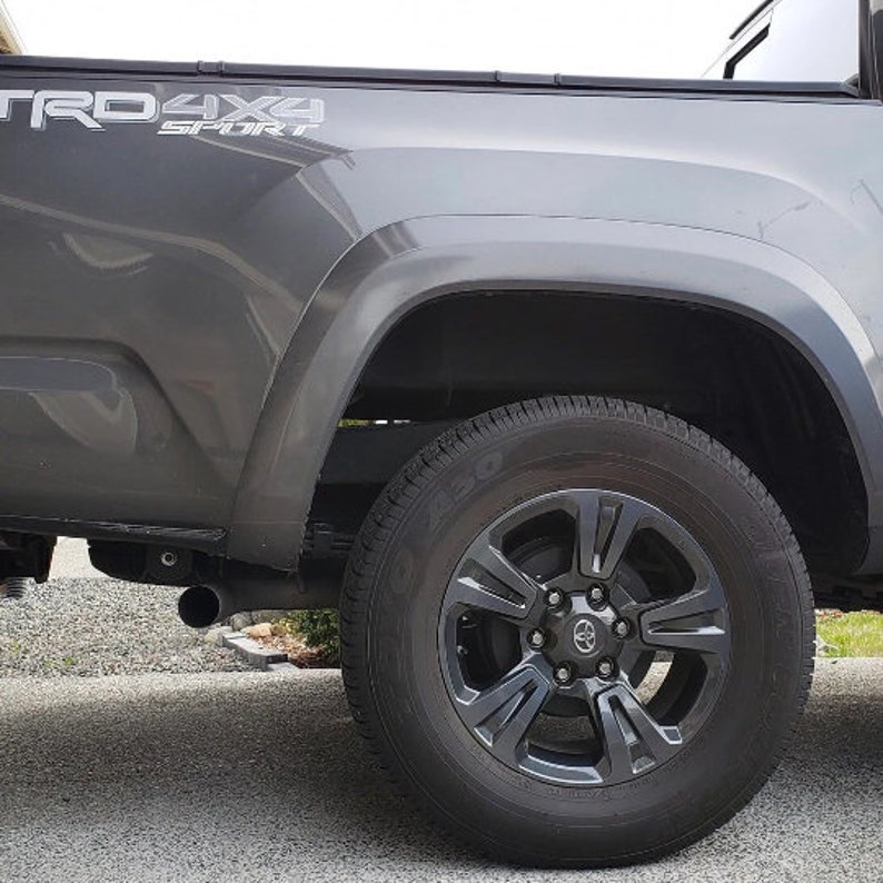 Może przedstawiać: Zbliżenie boku szarej ciężar&oacute;wki Toyota Tacoma. Opona jest czarna z ciemnoszarą felgą. Tekst "TRD 4x4 SPORT" jest widoczny z boku ciężar&oacute;wki.
