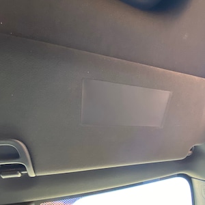 May include: A black car sun visor, fixed to the vehicle's roof. The visor features a rectangular shape with a textured surface and a rectangular mirror. A black plastic clip is visible.