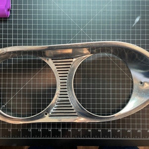 May include: A chrome-finished headlight bezel with two circular openings and a central, slotted section. The bezel has a streamlined, vintage design, likely for a classic car. The item is set against a black grid background.