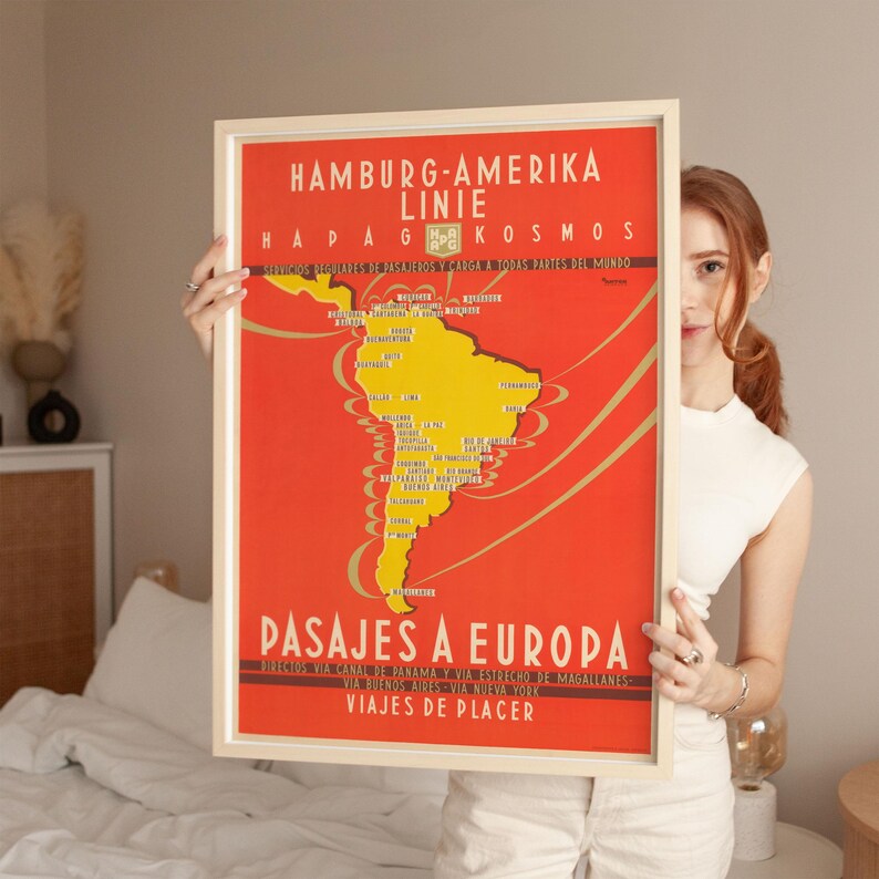 Op de afbeelding: Een ingelijste vintage reisposter met een rode achtergrond en een gele kaart van Zuid-Amerika. De poster heeft tekst in het Duits en Spaans, waaronder "Hamburg-Amerika Linie" en "Pasajes a Europa."