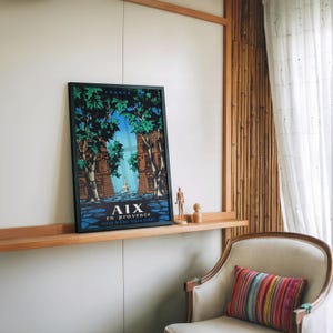 May include: A framed travel poster for Aix-en-Provence, France, with a colourful illustration of a street scene. The poster is displayed on a wooden shelf with small wooden figurines. A striped pillow rests on a cream-coloured armchair.