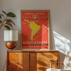 Op de afbeelding: Een ingelijste vintage reisposter met een rode achtergrond en een gele kaart van Zuid-Amerika. De poster heeft tekst in het Duits en Spaans en adverteert cruises naar Europa. De poster wordt weergegeven op een houten kast.