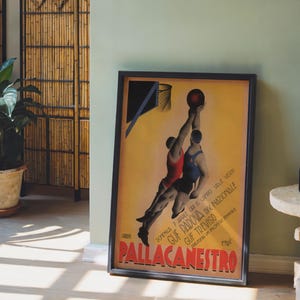 May include: Framed vintage basketball poster with two players reaching for the ball. The poster has a yellow background with text in Italian. The poster is in a black frame and leans against a light green wall.