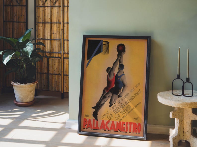 May include: Framed vintage basketball poster with two players reaching for the ball. The poster has a yellow background with text in Italian. The poster is in a black frame and leans against a light green wall.