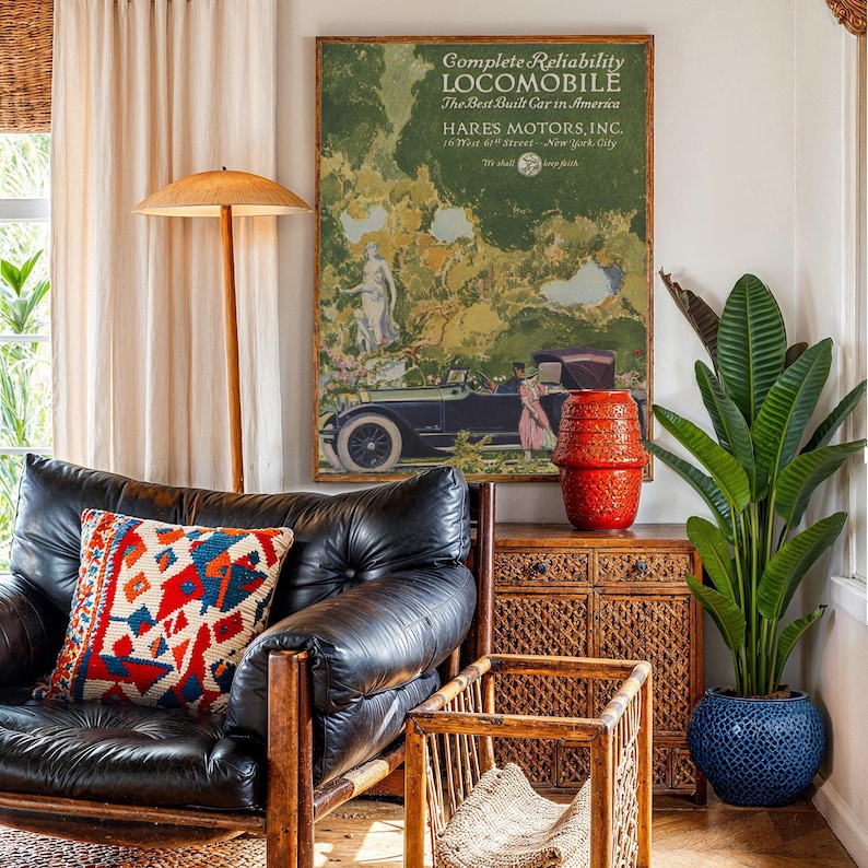 May include: An interior scene featuring a black leather armchair with a colorful patterned pillow. A vintage advertisement for a "LOCOMOBILE" car hangs on the wall. A red textured vase sits atop a woven cabinet, and a large potted plant adds greenery.