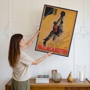 May include: A framed vintage basketball poster with Italian text, featuring two players in red and blue uniforms reaching for a basketball. The poster has a yellow background and a black frame. The poster is being held up.
