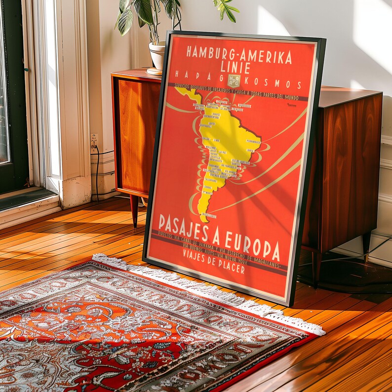 Op de afbeelding: Een ingelijste vintage reisposter met een rode achtergrond en een gele kaart van Zuid-Amerika. De poster heeft tekst in het Duits en Spaans en adverteert cruises. De poster leunt tegen een houten kast.