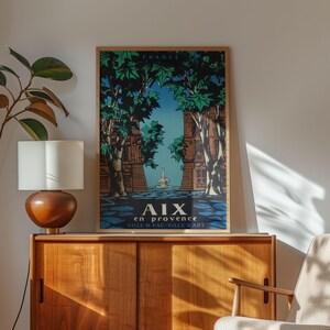 May include: A framed vintage travel poster for Aix-en-Provence, France. The artwork depicts a street scene with tall trees, buildings, and a fountain, in blue, green, and brown. The poster is displayed on a wooden cabinet.