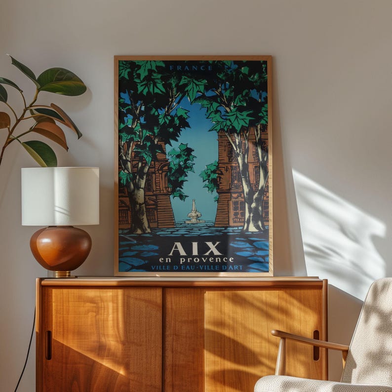 May include: A framed vintage travel poster for Aix-en-Provence, France. The artwork depicts a street scene with tall trees, buildings, and a fountain, in blue, green, and brown. The poster is displayed on a wooden cabinet.