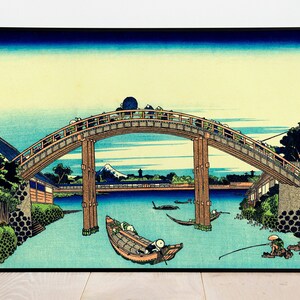 May include: A woodblock print of a Japanese bridge over a river with boats and people on it. The bridge is arched and has a wooden railing. The river is blue and the sky is a light blue. There are mountains in the background.