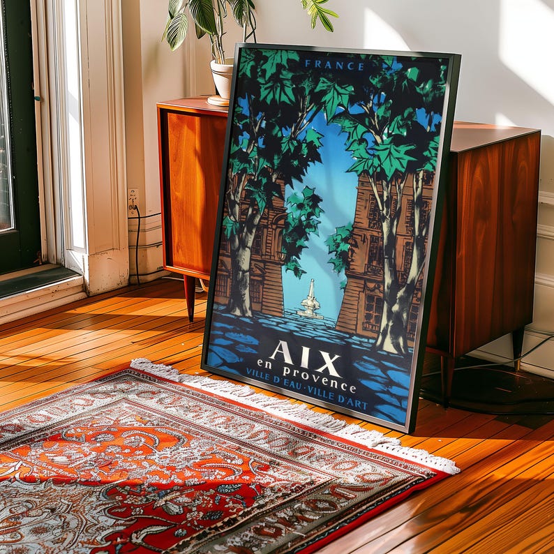 May include: Framed vintage travel poster for Aix-en-Provence, France. The poster features a blue sky, green trees, and architectural elements. The poster is in a black frame and leans against a wooden cabinet on a hardwood floor.