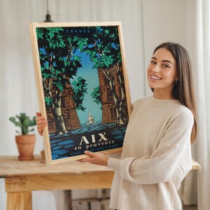 May include: A framed travel poster for Aix-en-Provence, France, with a light wood frame. The poster depicts a stylized illustration of buildings and trees, with the text "AIX en provence VILLE D'ART". The artwork uses green, blue, and brown tones.