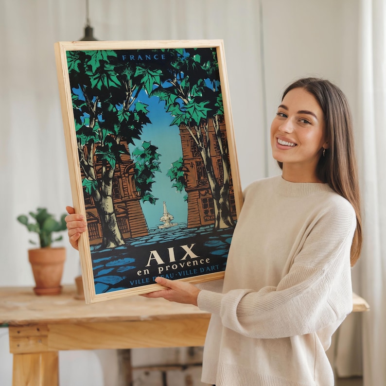 May include: A framed travel poster for Aix-en-Provence, France, with a light wood frame. The poster depicts a stylized illustration of buildings and trees, with the text "AIX en provence VILLE D'ART". The artwork uses green, blue, and brown tones.