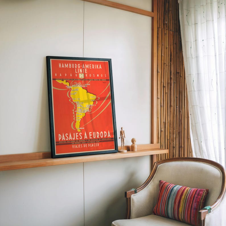Op de afbeelding: Een ingelijste vintage reisposter met een rode achtergrond en een gele kaart van Zuid-Amerika. De poster heeft zwarte tekst in het Duits en Spaans, die reizen naar Europa adverteert. De poster wordt op een houten plank tentoongesteld.
