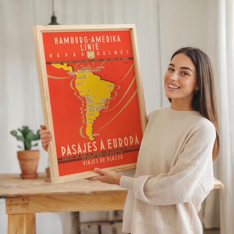 Op de afbeelding: Een ingelijste vintage reisposter met een rode achtergrond en een gele kaart van Zuid-Amerika. De poster heeft tekst in het Duits en Spaans, waaronder "Hamburg-Amerika Linie" en "Pasajes a Europa". De lijst is van lichtgekleurd hout.