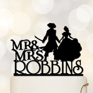May include: Black silhouette of a bride and groom wedding cake topper. The groom is holding a sword and the bride is wearing a gown. The text "MR & MRS ROBBINS" is below the couple.