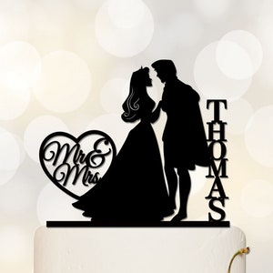 Puede incluir: Adorno para tarta con silueta negra que representa a una novia y un novio. El novio lleva una capa. Las palabras "Mr & Mrs" están dentro de un corazón, y el nombre "Thomas" se muestra verticalmente. La parte inferior del adorno dice "DESIGNED BY ACRYLICDESIGNFORYOU".