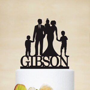 May include: Black silhouette cake topper of a family of four, including a couple, a baby, and a child. The topper is on a white cake with peach flowers. The text "GIBSON" is below the silhouettes.