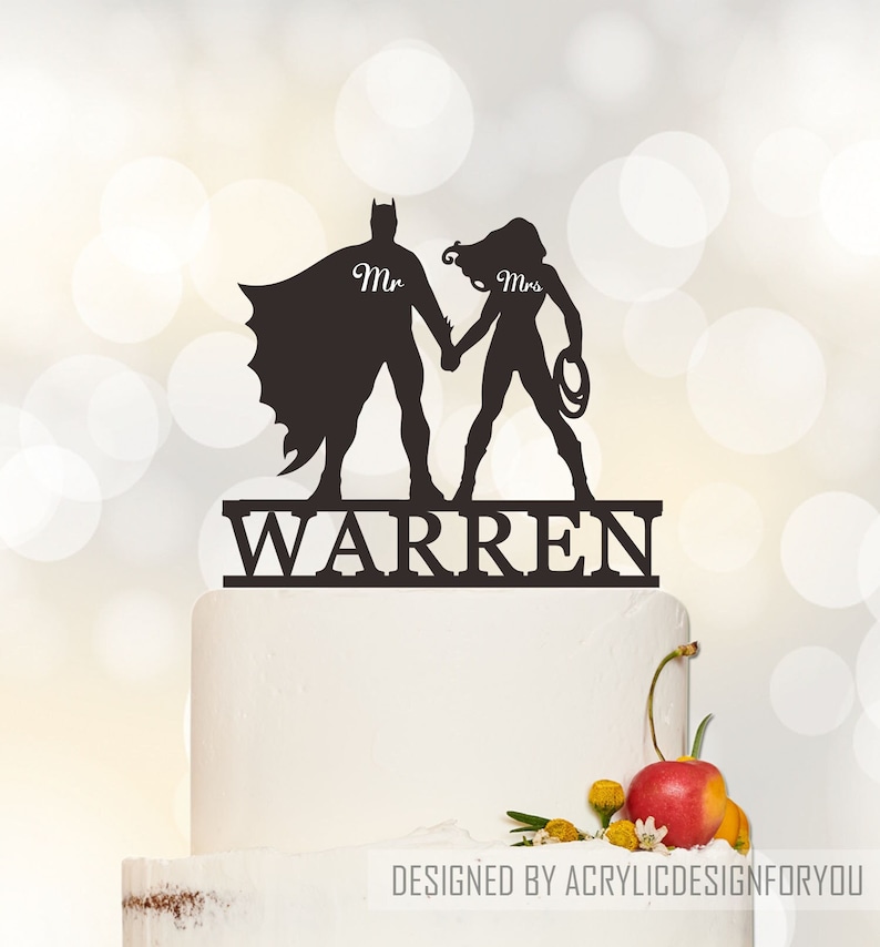May include: Black silhouette cake topper featuring Batman and Wonder Woman holding hands with the text "Mr." and "Mrs." above their heads and the name "WARREN" below.