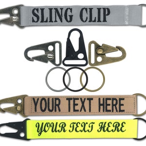 May include: Three keychains with different colored straps and metal clips. The top keychain has a gray strap with the text "SLING CLIP" in black. The middle keychain has a tan strap with the text "YOUR TEXT HERE" in black. The bottom keychain has a yellow strap with the text "YOUR TEXT HERE" in black.