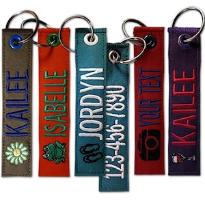 May include: A collection of personalized fabric keychains in various colors, each with embroidered text and designs. The keychains feature names, a phone number, and icons like a camera, wine glass, and a frog.