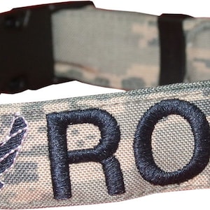 May include: A camouflage dog collar with a black embroidered name "ROCK" and a silver Air Force wing patch.