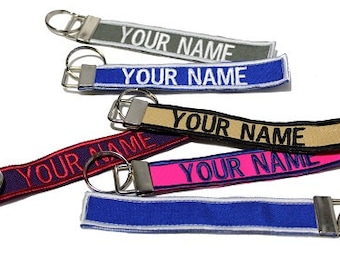 Op de afbeelding: Vijf verschillende sleutelhangers met metalen ringen en stoffen banden. De banden zijn geborduurd met de tekst "YOUR NAME" in verschillende kleuren, waaronder rood, roze, blauw, zwart en grijs.