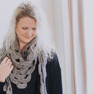 Peut inclure: Une femme portant une &eacute;charpe en crochet gris avec un pull noir. L'&eacute;charpe a un motif en dentelle et est drap&eacute; autour de son cou.