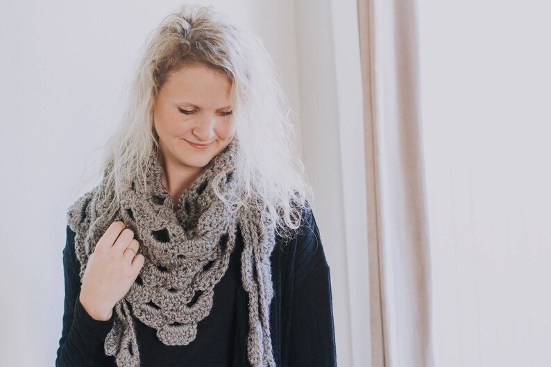 Peut inclure: Une femme portant une &eacute;charpe en crochet gris avec un pull noir. L'&eacute;charpe a un motif en dentelle et est drap&eacute; autour de son cou.