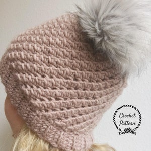 May include: A pink crocheted beanie with a fluffy gray pom pom on top. The beanie has a textured stitch pattern and is made with a soft yarn. Crochet Pattern.
