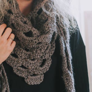 Peut inclure: Gros plan d'une personne portant une &eacute;charpe en crochet marron avec un motif en dentelle. L'&eacute;charpe est drap&eacute; autour du cou et des &eacute;paules de la personne.
