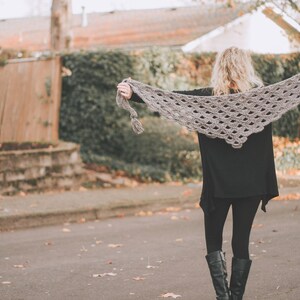 Peut inclure: Une femme portant une &eacute;charpe en crochet grise avec un bord festonn&eacute;. Elle se tient devant une cl&ocirc;ture en bois et de la v&eacute;g&eacute;tation verte.
