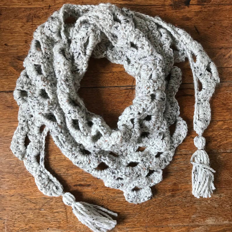 Peut inclure: Une &eacute;charpe en crochet blanche avec un motif ajour&eacute; et deux pompons. L'&eacute;charpe est pos&eacute;e sur une surface en bois.