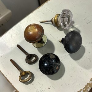 Puede incluir: Una colección de herrajes que incluye un pomo de cristal transparente facetado con base de latón, un pomo redondo de cobre deslustrado, un pomo redondo negro mate, un pomo redondo oscuro moteado y dos cáncamos de latón.