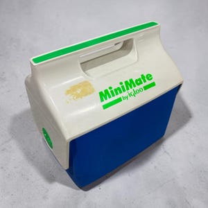 May include: A white and blue plastic portable cooler with a top handle. The lid features the green text MiniMate by igloo and a green accent strip. There is a small area of yellow discoloration on the white lid surface near the handle.