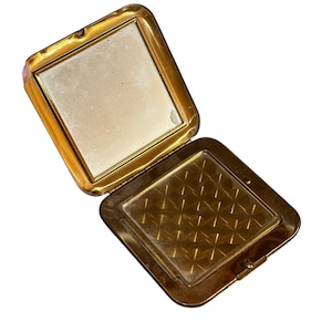 May include: A square gold-toned metal compact case with a hinged lid. The interior features a square mirror on the top half and a square powder compartment on the bottom half, which is covered by a clear plastic insert with a geometric grid pattern.