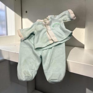 May include: A light mint green two-piece outfit consisting of a long-sleeved top and footed trousers. The top features a white vertical centre panel, white sleeve cuffs, and a small embroidered green arch design on the upper left chest area.