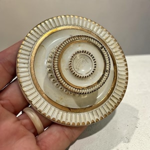 May include: A round, off-white ceramic lid featuring a raised, tiered center knob with a beaded texture. The lid is decorated with gold-painted trim along the outer fluted edge and the central circular rings, creating a layered, circular design.