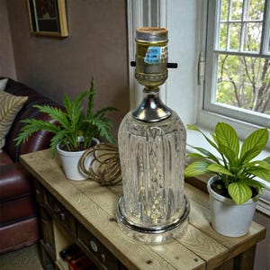 Puede incluir: Una lámpara de mesa vintage con una base de vidrio tallado transparente y una parte superior de metal plateado. La lámpara tiene forma cilíndrica con ranuras verticales y un portalámparas de metal. La lámpara está sobre una mesa de madera con plantas.