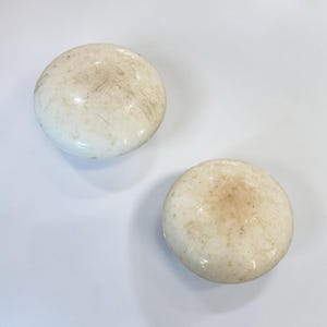May include: Two round, off-white ceramic knobs with a smooth, glossy finish. Each knob features a slightly domed shape and shows subtle, light brown surface markings and natural variations in the glaze across the circular tops.