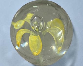 May include: A spherical clear glass paperweight featuring an internal design of a yellow flower with delicate petals suspended within the transparent material. The glass orb has a smooth, polished surface that reflects light across its rounded form.