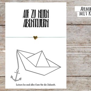Könnte beinhalten: Eine weiße Karte mit dem Text "Auf zu neuen Abenteuern" und einem kleinen goldenen Herz-Anhänger. Unter dem Text befindet sich ein graues Papierboot mit einem grauen Anker, der daran befestigt ist. Der Text "Leinen los und alles Gute für die Zukunft" ist unter dem Boot gedruckt. Die Karte ist mit "Armband mit Karte" beschriftet.