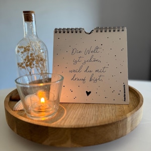 Könnte beinhalten: Eine weiße Karte mit schwarzem Text und einem kleinen schwarzen Herz lautet "Die Welt ist schön, weil du mit drauf bist." Die Karte liegt auf einem Holztablett mit einem durchsichtigen Glas-Kerzenhalter und einer mit getrockneten Blumen gefüllten Flasche.