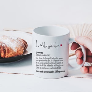 May include: White ceramic mug with a black and red design. The mug says "Lieblingskollege" and includes a definition of the word "Jonas" in German. The mug is held by a hand.