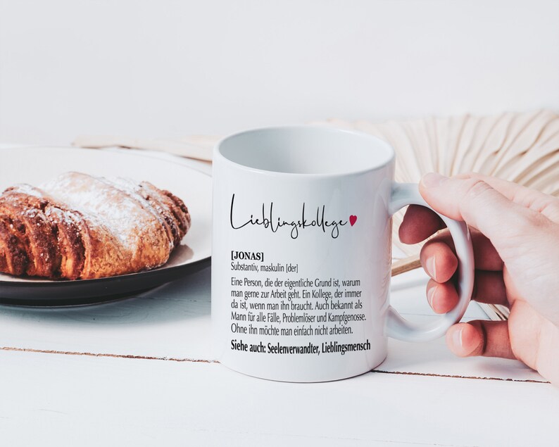 May include: White ceramic mug with a black and red design. The mug says "Lieblingskollege" and includes a definition of the word "Jonas" in German. The mug is held by a hand.