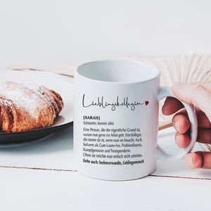 May include: White ceramic mug with a black and red design. The mug has the text "Lieblingskollegin" and a definition of the word "Sarah" in German. The mug is being held by a hand.