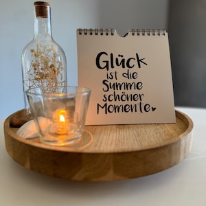 Könnte beinhalten: Ein Holztablett mit einem kleinen Glas-Kerzenhalter, einer durchsichtigen Glasflasche mit getrockneten Blumen und einer weißen Karte mit dem Text "Glück ist die Summe schöner Momente" in schwarzer Schrift.