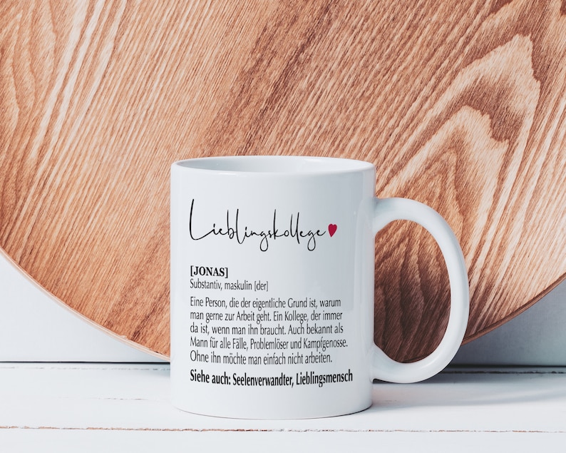 May include: White ceramic mug with a black and red heart design. The mug has a black text that reads "Lieblingskollege [JONAS] Substantiv, maskulin (der) Eine Person, die der eigentliche Grund ist, warum man gerne zur Arbeit geht. Ein Kollege, der immer da ist, wenn man ihn braucht. Auch bekannt als Mann f&uuml;r alle F&auml;lle, Probleml&ouml;ser und Kampfgenosse. Ohne ihn m&ouml;chte man einfach nicht arbeiten. Siehe auch: Seelenverwandter, Lieblingsmensch"